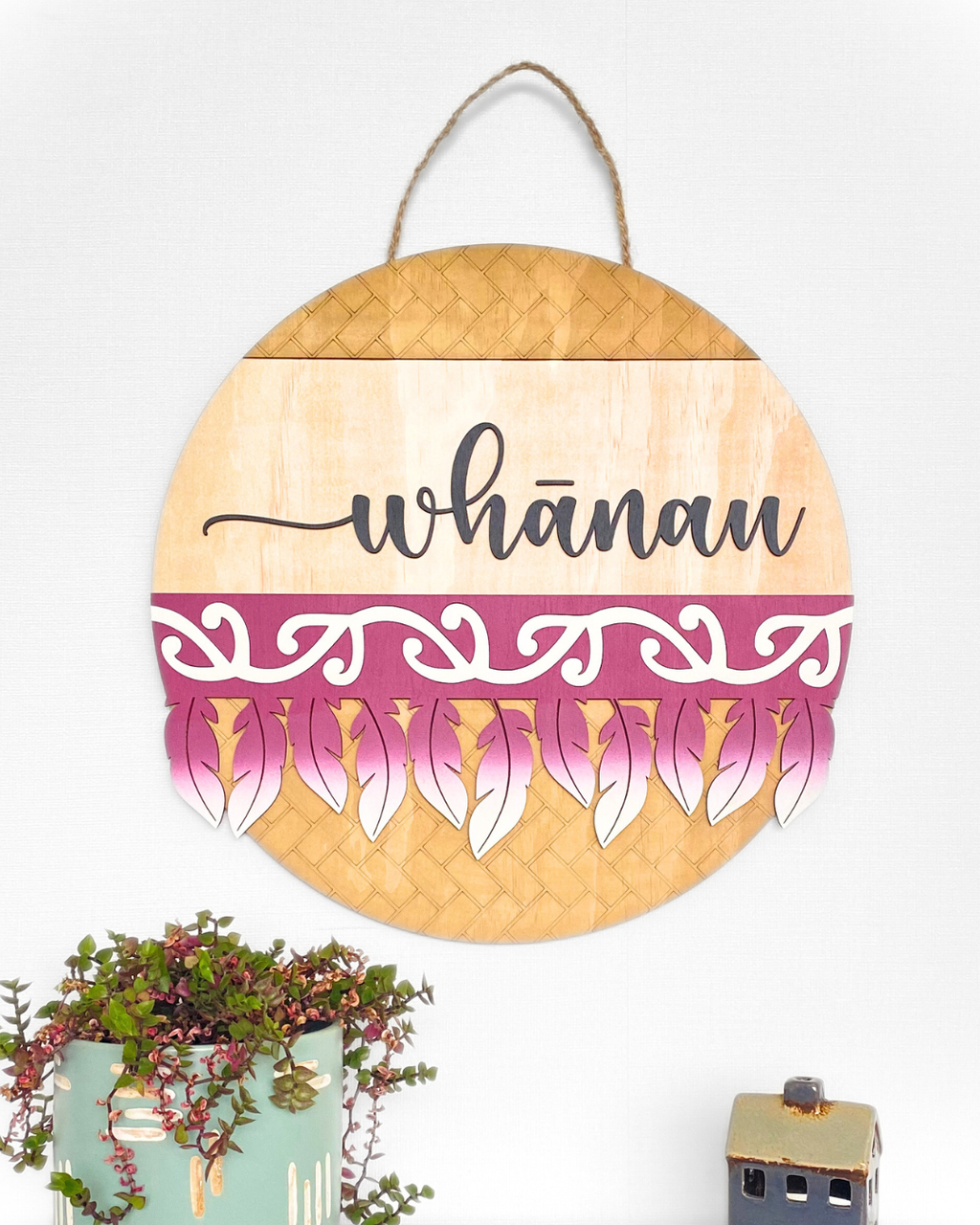 The Feather Round wall art complete with the word 'whānau' in a script font, made from natural plywood and hand-painted with a traditional Māori pattern or motif, in the colour Burgundy.