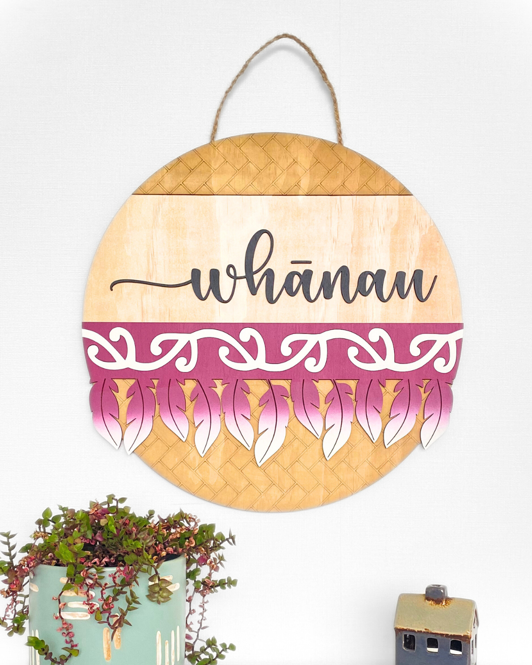 The Feather Round wall art complete with the word 'whānau' in a script font, made from natural plywood and hand-painted with a traditional Māori pattern or motif, in the colour Burgundy.