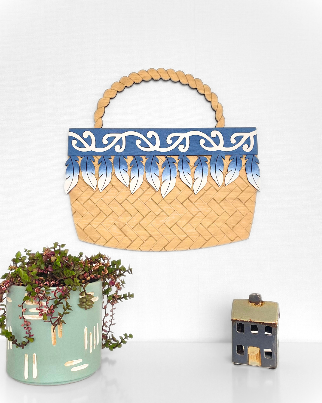 The Feather Kete wall art, made from natural plywood and hand-painted in the shape of a woven basket with a traditional Māori pattern or motif, in the colour Denim Blue.