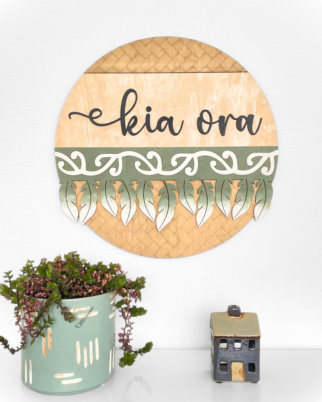 The Feather Round wall art complete with the worda 'kia ora' in a script font, made from natural plywood and hand-painted with a traditional Māori pattern or motif, in the colour Olive Green.