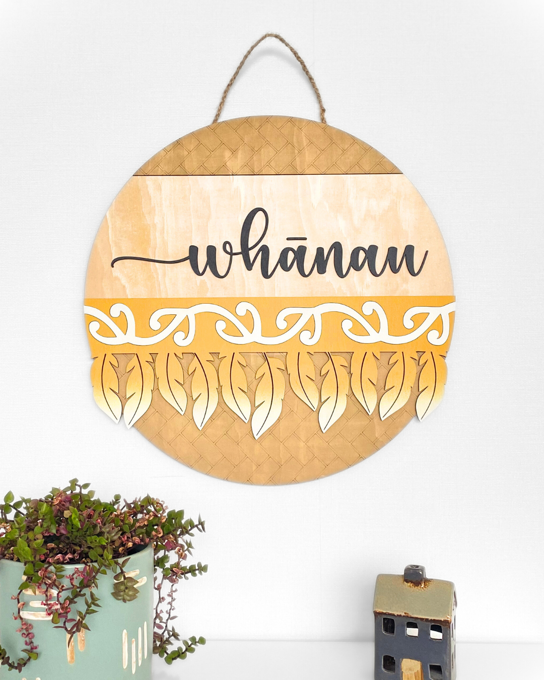The Feather Round wall art complete with the word 'whānau' in a script font, made from natural plywood and hand-painted with a traditional Māori pattern or motif, in the colour Mustard.