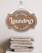 Wooden sign with 'Self Service Laundry Open 24 Hours' text above folded towels on a white background