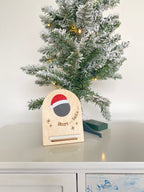 Wooden countdown calendar placed next to a mini Christmas tree, with mini chalkboard and glittery red Santa hat. Comes with chalk.