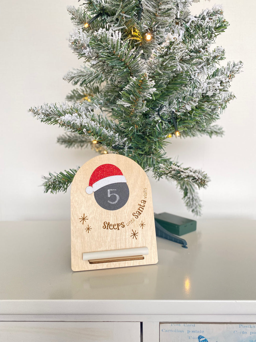 Wooden countdown calendar placed next to a mini Christmas tree, with mini chalkboard and glittery red Santa hat. Comes with chalk.