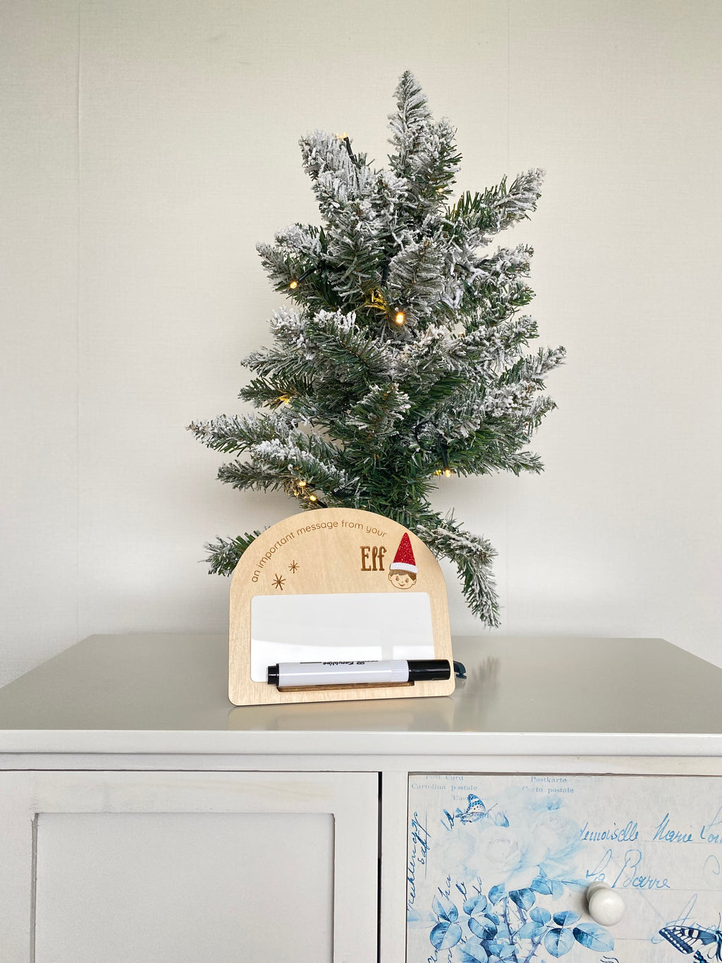 Small wooden plaque next to a mini Xmas tree, with a mini whiteboard and marker, decorated with Christmas and Elf on the Shelf motifs.