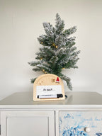 Small wooden plaque next to a mini Xmas tree, with a mini whiteboard and marker, decorated with Christmas and Elf on the Shelf motifs. The whiteboard has the words 'I'm back' written on it by the elf.