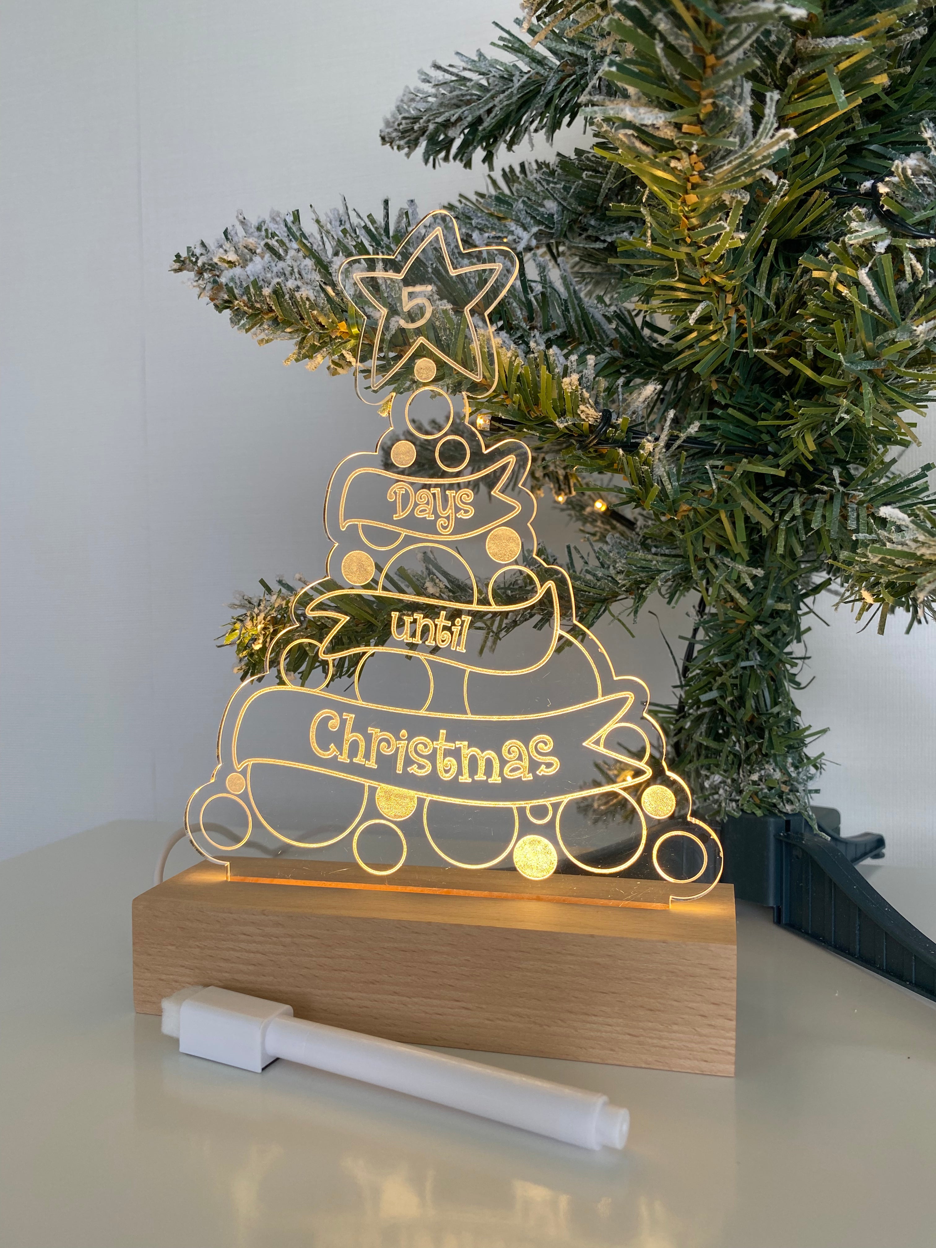 Christmas Countdown Night Light placed next to a mini Xmas tree, made from clear acrylic on a wooden base, complete with a whiteboard marker to write down each day of advent. Powered by USB.